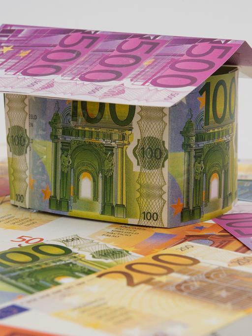 Ein Haus aus künstlichen Geldscheinen steht  auf weiteren Geldscheinen
