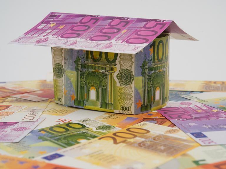 Ein Haus aus künstlichen Geldscheinen steht  auf weiteren Geldscheinen