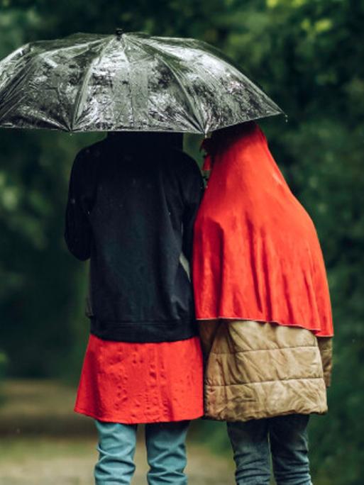 Zwei Menschen sind in Regenjacken eingehüllt und stehen unter einem Regenschirm, damit sie sich gegen Unwetter schützen.