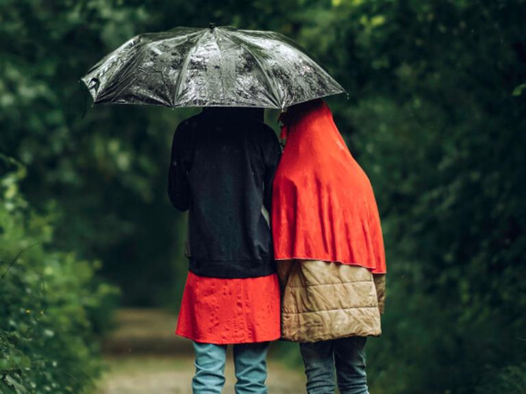 Zwei Menschen sind in Regenjacken eingehüllt und stehen unter einem Regenschirm, damit sie sich gegen Unwetter schützen.