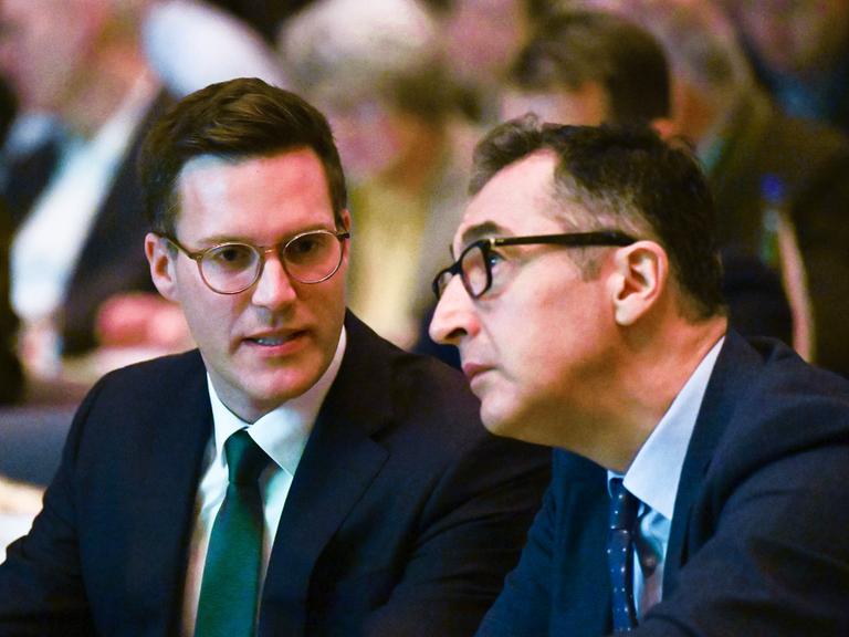 Manuel Hagel (l, CDU) und Cem Özdemir (r, Bündnis 90 / Die Grünen) sitzen nebeneinander bei einer Veranstaltung. Ihre Köpfe sind einander zugeneigt, sie scheinen im Gespräch.