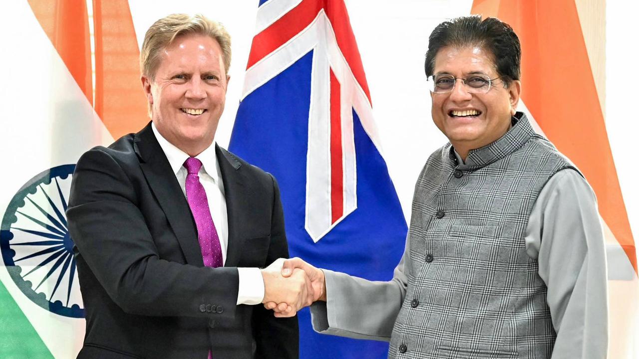 Todd McClay und Piyush Goyal geben sich die Hand und blicken Richtung Kamera. Im Hintergrund die Flaggen Neuseelands und Indiens