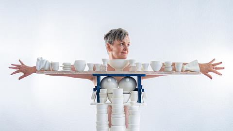 Die Künstlerin Uli Aigner steht hinter einem Gestell mit vielen weiße Porzellanobjekten aus ihrem Kunstprojekt "One Million", und breitet die Arme aus. Das Gestell mit den Porzellanteilen formt ihren Körper nach. 