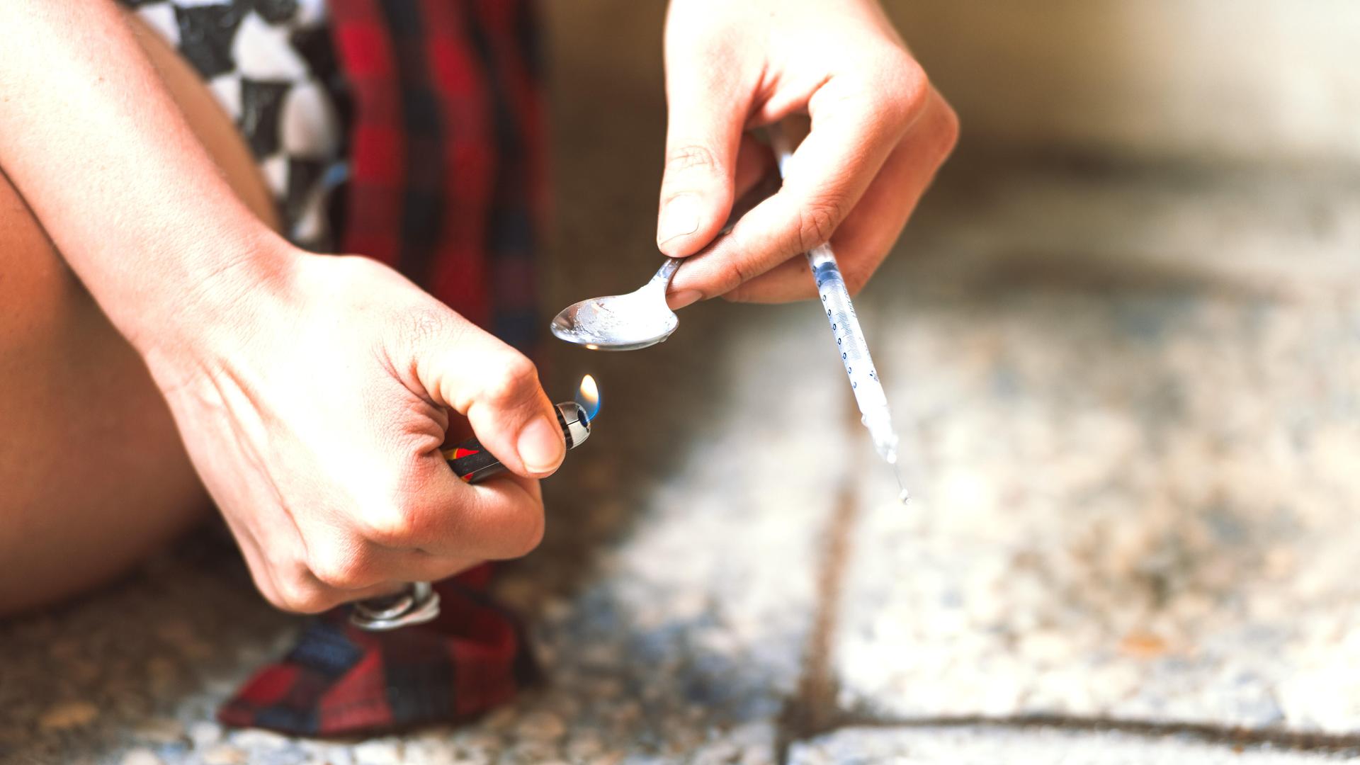 Zwei Hände halten ein brennendes Feuerzeug und einen Löffel mit einer Spritze