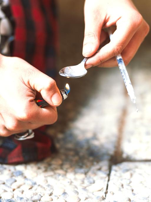 Zwei Hände halten ein brennendes Feuerzeug und einen Löffel mit einer Spritze