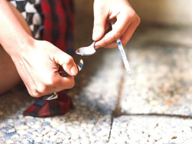 Zwei Hände halten ein brennendes Feuerzeug und einen Löffel mit einer Spritze