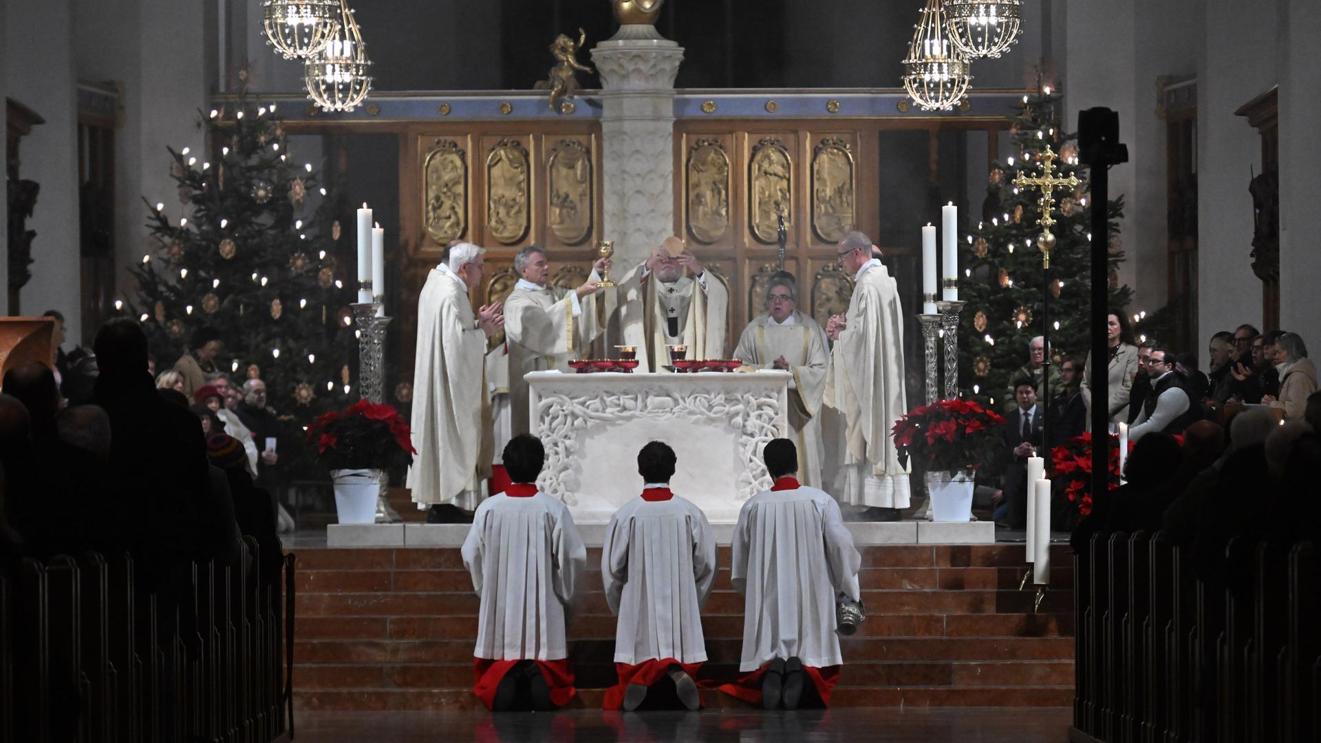 Marx feiert die Heilige Messe. Er hält dabei neben einem Kelch eine Hostie in die Höhe. Die Kirche ist weihnachtlich geschmückt. 