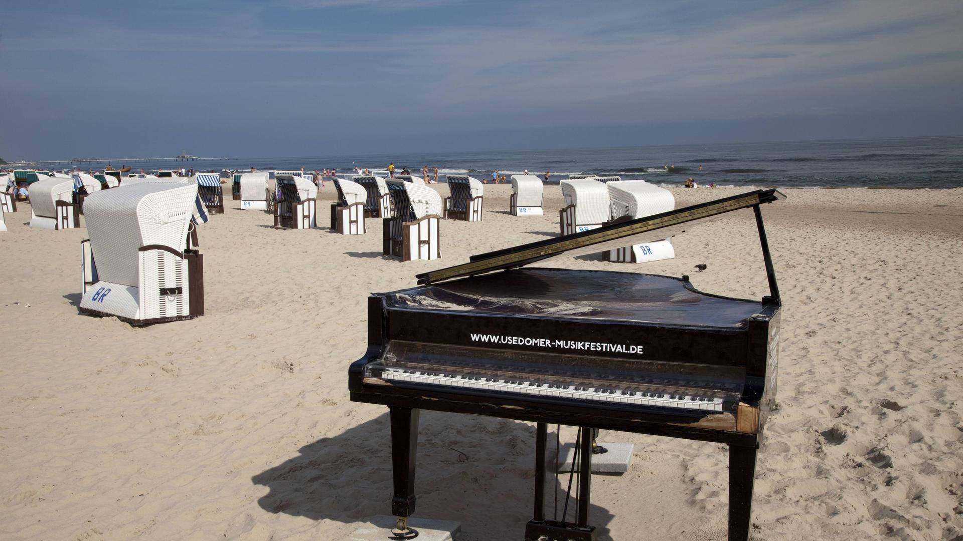 Wir sehen einen Klavier-Flügel am Strand, der vor vielen Strandkörben steht. Im Hintergrund das Meer. Wir sehen einen Klavier-Flügel am Strand, der vor vielen Strandkörben steht. Im Hintergrund das Meer.