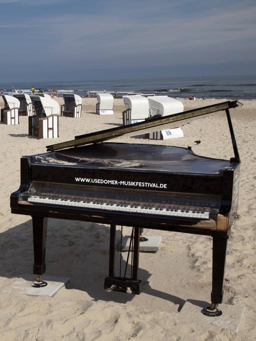 Wir sehen einen Klavier-Flügel am Strand, der vor vielen Strandkörben steht. Im Hintergrund das Meer. 