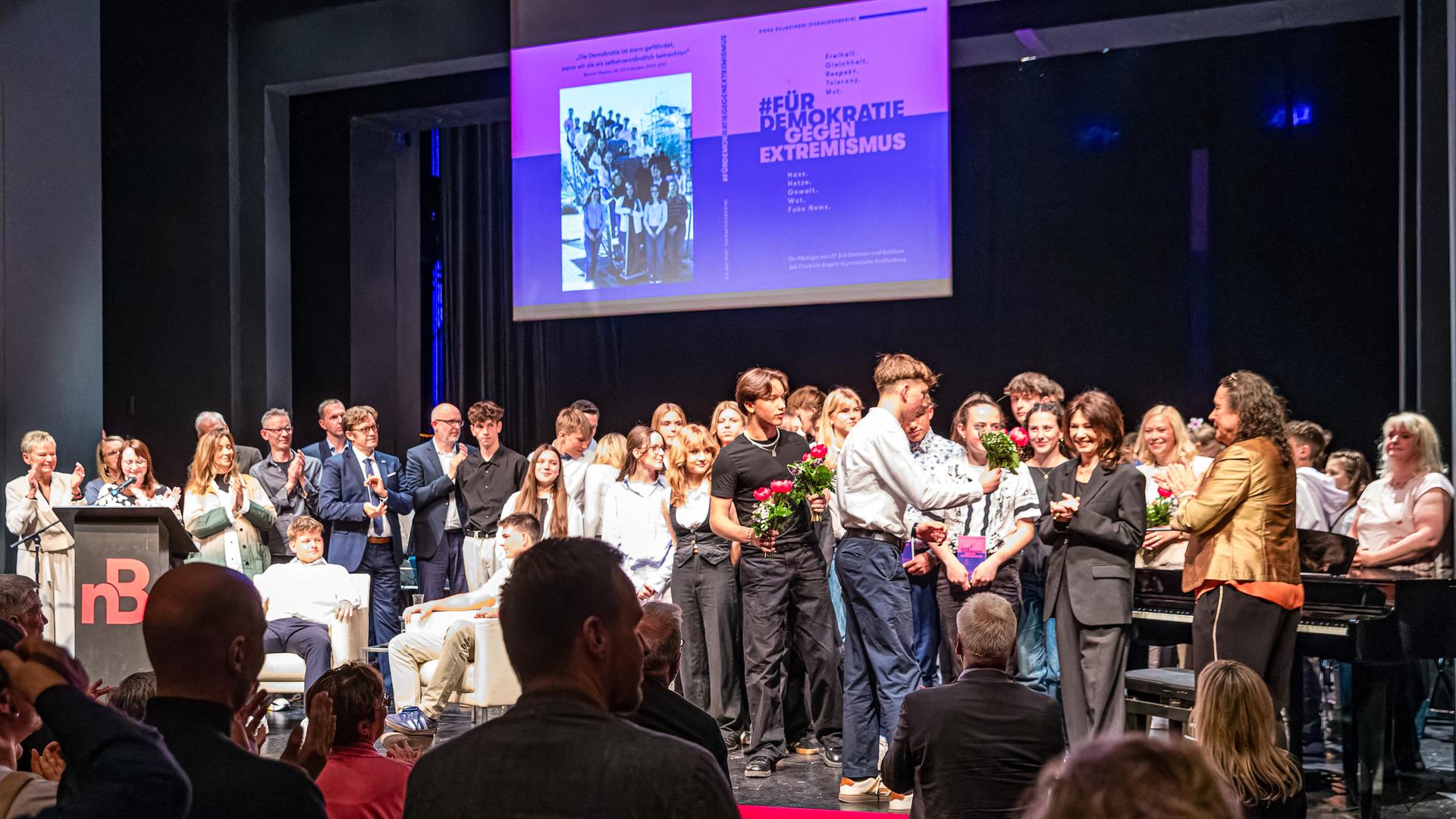 Autorinnen, Autoren und andere Beteiligte stehen bei der Premiere für das Buch "#FürDemokratieGegenExtremismus" auf der Bühne im Theater Senftenberg. Iris Berben (6.v.r.), Schauspielerin, hatte ein Vorwort verfasst und erhält einen Blumenstrauß.