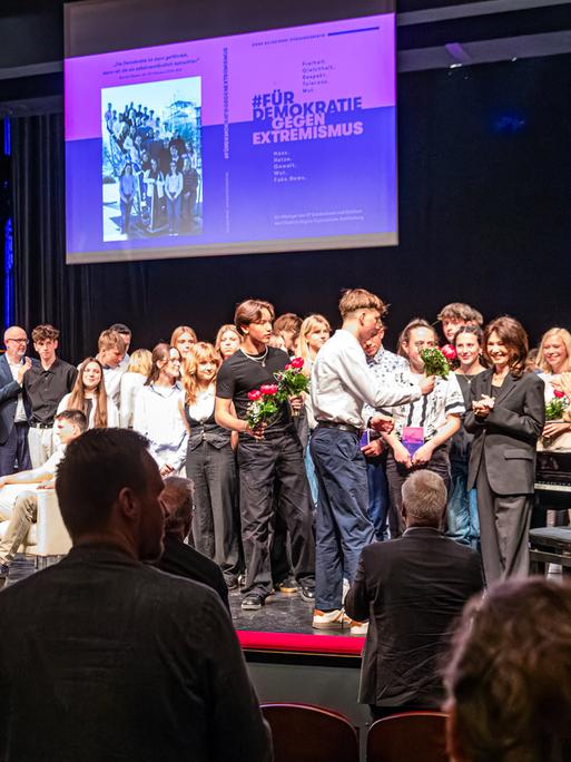 Autorinnen, Autoren und andere Beteiligte stehen bei der Premiere für das Buch "#FürDemokratieGegenExtremismus" auf der Bühne im Theater Senftenberg. Iris Berben (6.v.r.), Schauspielerin, hatte ein Vorwort verfasst und erhält einen Blumenstrauß.
