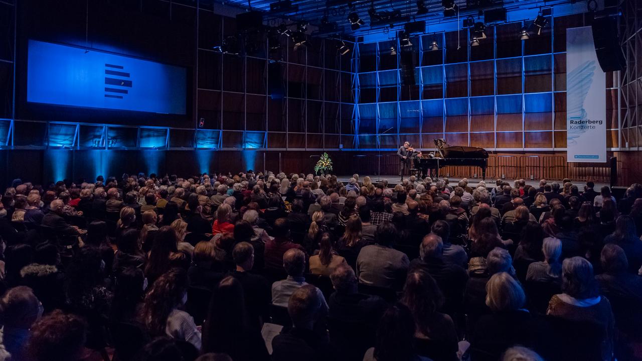 Ein Geiger und ein Pianist geben ein Konzert in einem voll besetzten, holzvertäfelten Saal, der blau beleuchtet ist.