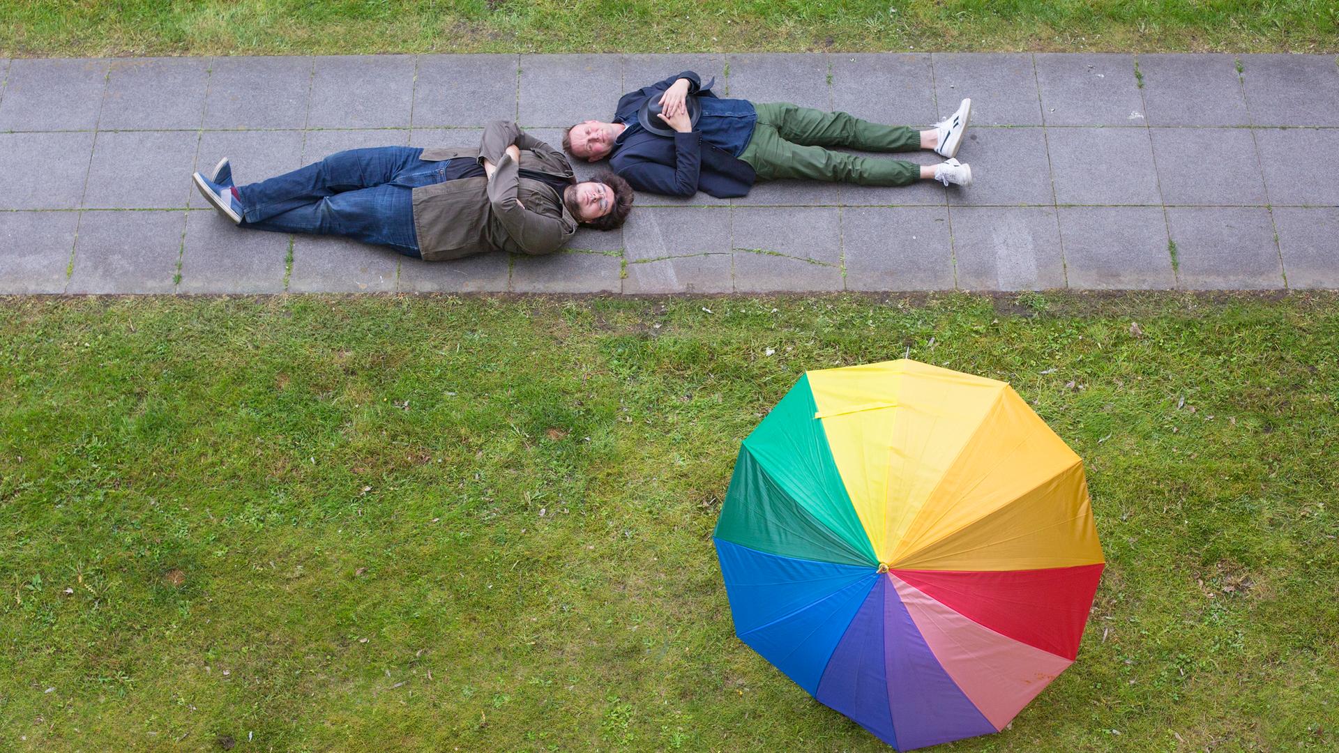 Devid Striesow und Axel Rahnisch liegen rücklings auf einem Fußgängerweg durch eine grüne Wiese,  auf der widerum ein regenbogenfarbener Schirm liegt.