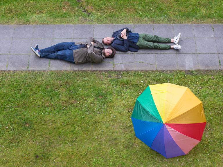 Devid Striesow und Axel Rahnisch liegen rücklings auf einem Fußgängerweg durch eine grüne Wiese,  auf der widerum ein regenbogenfarbener Schirm liegt.
