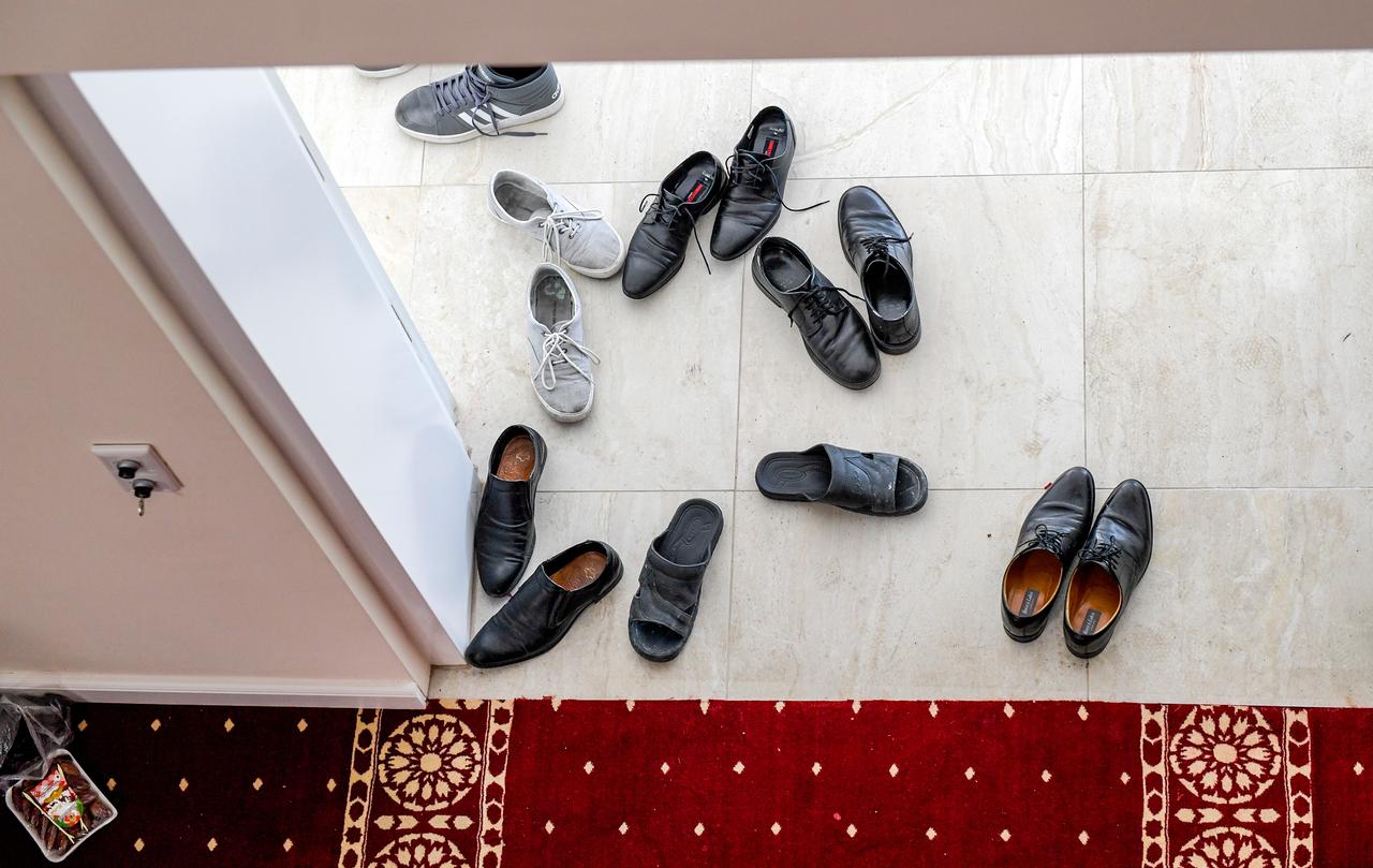 Vor einem Türrahmen stehen Paare verschiedener Schuhe – Turnschuhe, schwarze Lederschuhe und Sandalen – vorn ist ein roter Teppich mit weißen Ornamenten zu sehen.