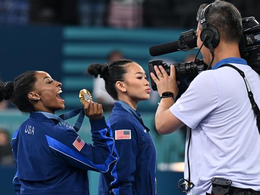 Olympia, Paris 2024, Turnen, Mehrkampf, Frauen, Finale, Simone Biles (l) aus den USA jubelt mit ihrer Goldmedaille vor einer TV-Kamera.