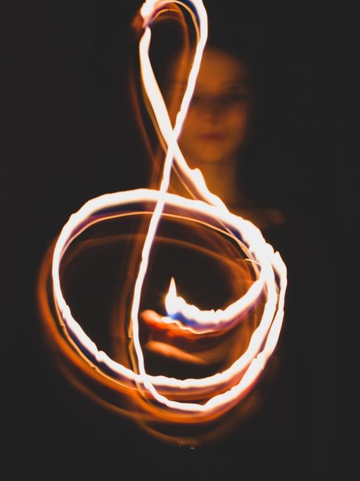 Eine junge Frau formt in der Dunkelheit mit einer Flamme einen Notenschlüssel, der durch eine Langzeitaufnahme sichtbar wird.