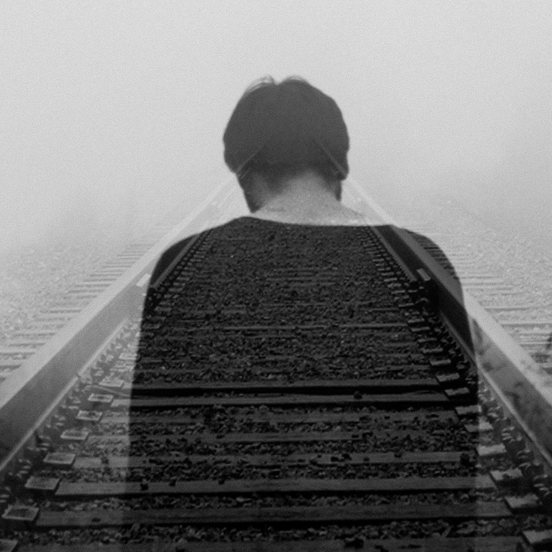 Schwarzweißfotografie eines Zuggleises, das im Nebel in in die Tiefe des Raumes führt. In einer Doppelbelichtung ist darüber die halbdurchsichtige Rückansicht eines Mannes zu sehen. Die Stimmung des Bildes ist sehr melancholisch.