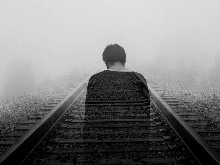 Schwarzweißfotografie eines Zuggleises, das im Nebel in in die Tiefe des Raumes führt. In einer Doppelbelichtung ist darüber die halbdurchsichtige Rückansicht eines Mannes zu sehen. Die Stimmung des Bildes ist sehr melancholisch.
