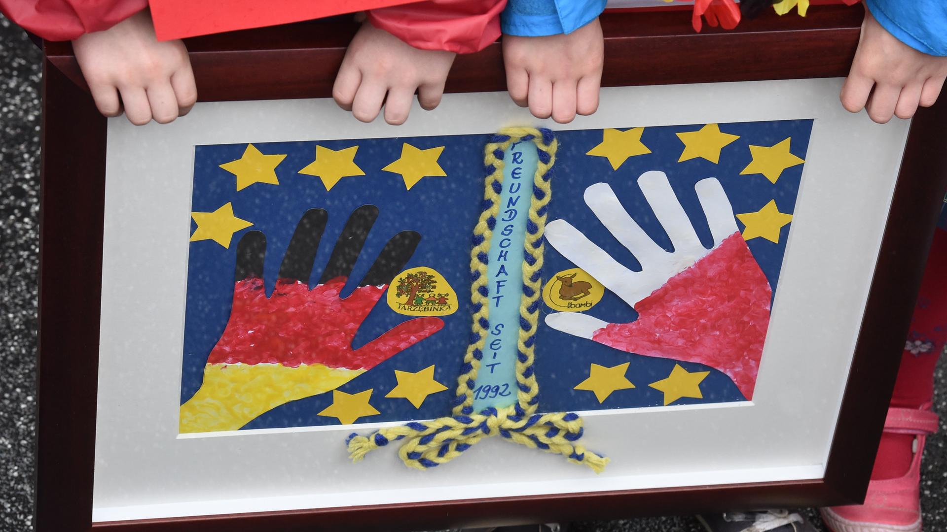 Zwei Kinder der Kita Bambi aus Frankfurt (Oder) halten ein selbst gemaltes Bild in einem Rahmen, das zwei Hände in den polnischen und deutschen Nationalfarben umringt von gelben Europasternen zeigt. 