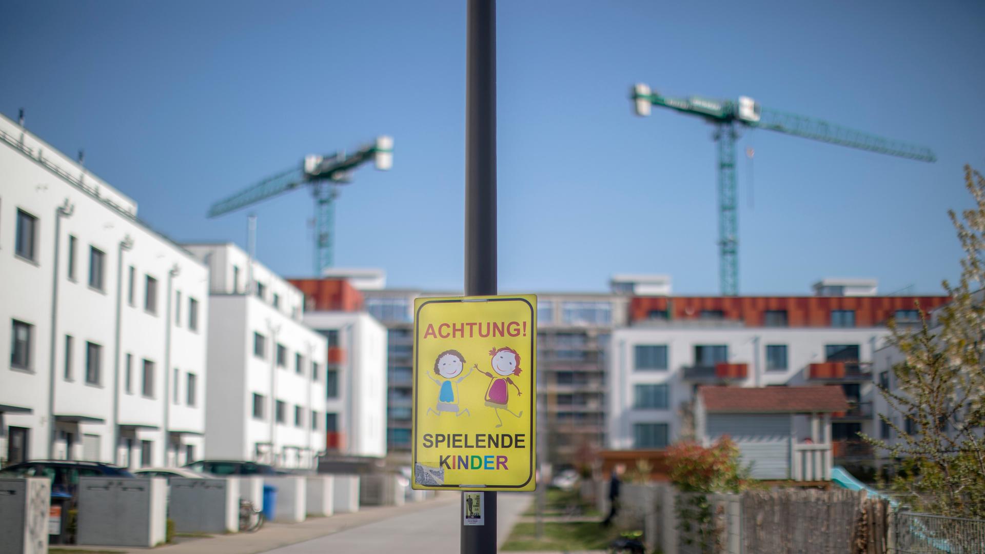 Neu errichtete Häuser stehen in einem Neubaugebiet in der Stadt Berlin. Ein Hinweisschild warnt vor spielenden Kindern, im Hintergrund ragen Baukräne in den Himmel