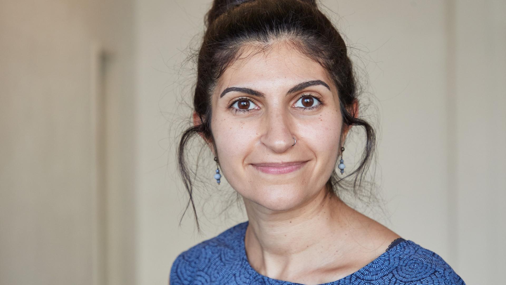 Shida Bazyar schaut in die Kamera. Sie hat ihre schwarzen Haare hochgesteckt, ihr Kleid ist blau-schwarz gemustert