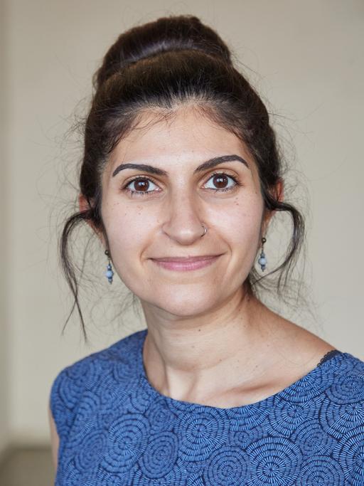 Shida Bazyar schaut in die Kamera. Sie hat ihre schwarzen Haare hochgesteckt, ihr Kleid ist blau-schwarz gemustert