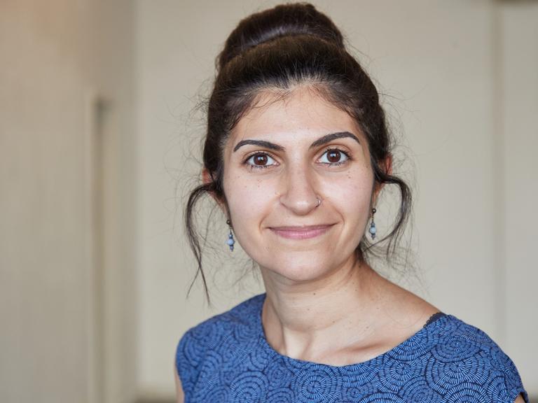 Shida Bazyar schaut in die Kamera. Sie hat ihre schwarzen Haare hochgesteckt, ihr Kleid ist blau-schwarz gemustert