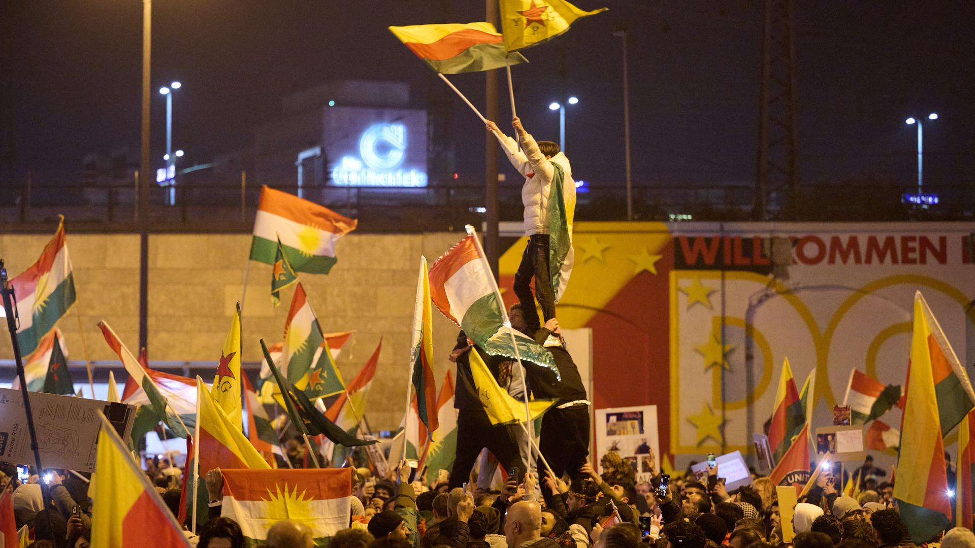 Menschen mit Fahnen und Plakaten stehen in gelblichem Laternenlicht.