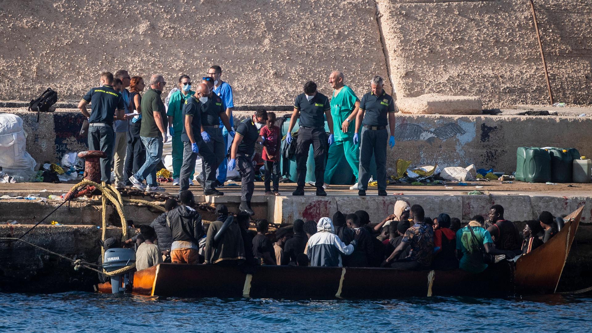Viele Menschen auf einem Boot kommen an einem Hafen in Italien an. Helfer an Land strecken ihnen die Hand entgegen.