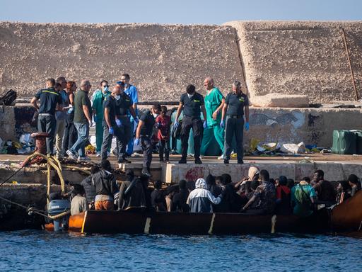 Viele Menschen auf einem Boot kommen an einem Hafen in Italien an. Helfer an Land strecken ihnen die Hand entgegen.