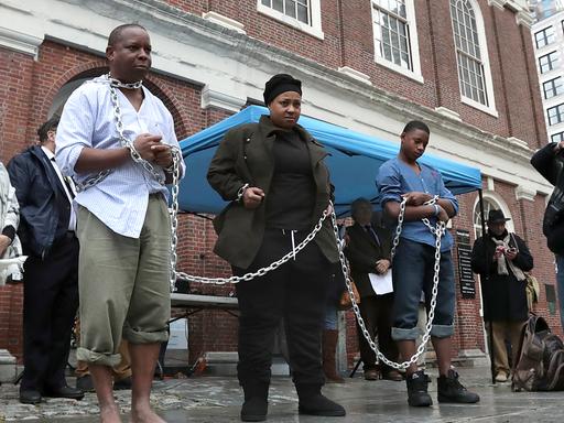 Eine Gruppe von PoC stehen mit Ketten nebeneinander gereiht vor einem Gebäude in Boston.