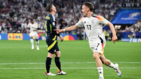 Florian Wirtz jubelt im DFB-Trikot; dahinter sind Spieler Schottlands und Deutschlands und voll besetzte Zuschauerränge zu erkennen.