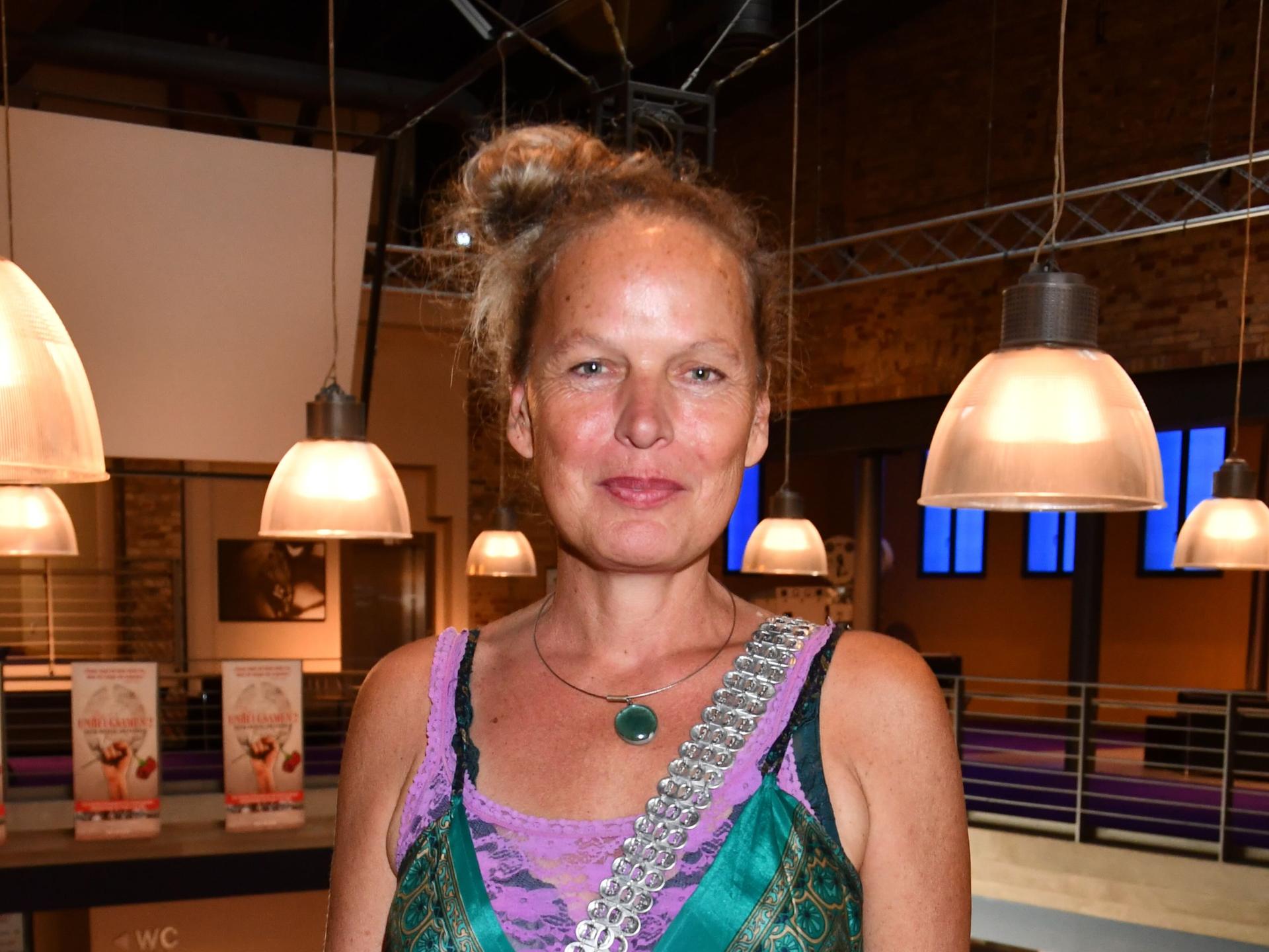 Amrei Bauer steht in einem Raum mit großen Lampenschirmen im Hintergrund. Sie trägt ein grünes Kleid und schaut fröhlich in die Kamera.