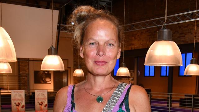 Amrei Bauer steht in einem Raum mit großen Lampenschirmen im Hintergrund. Sie trägt ein grünes Kleid und schaut fröhlich in die Kamera.
