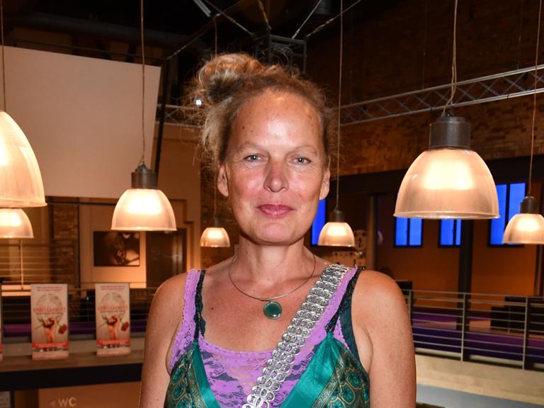 Amrei Bauer steht in einem Raum mit großen Lampenschirmen im Hintergrund. Sie trägt ein grünes Kleid und schaut fröhlich in die Kamera.