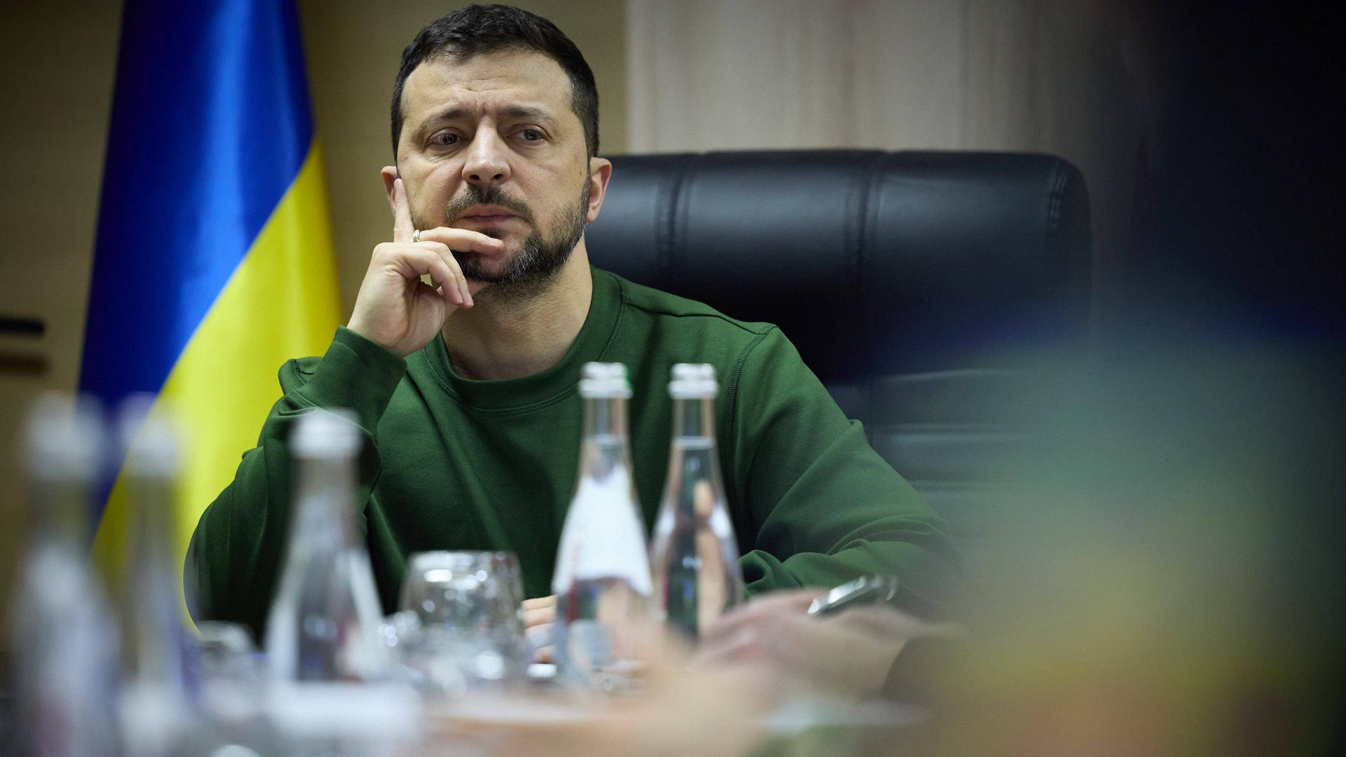 Wolodymyr Selenskyj sitzt an einem Tisch und macht ein nachdenkliches Gesicht. Im Hintergrund ist eine blau-gelbe Flagge seines Landes zu sehen.