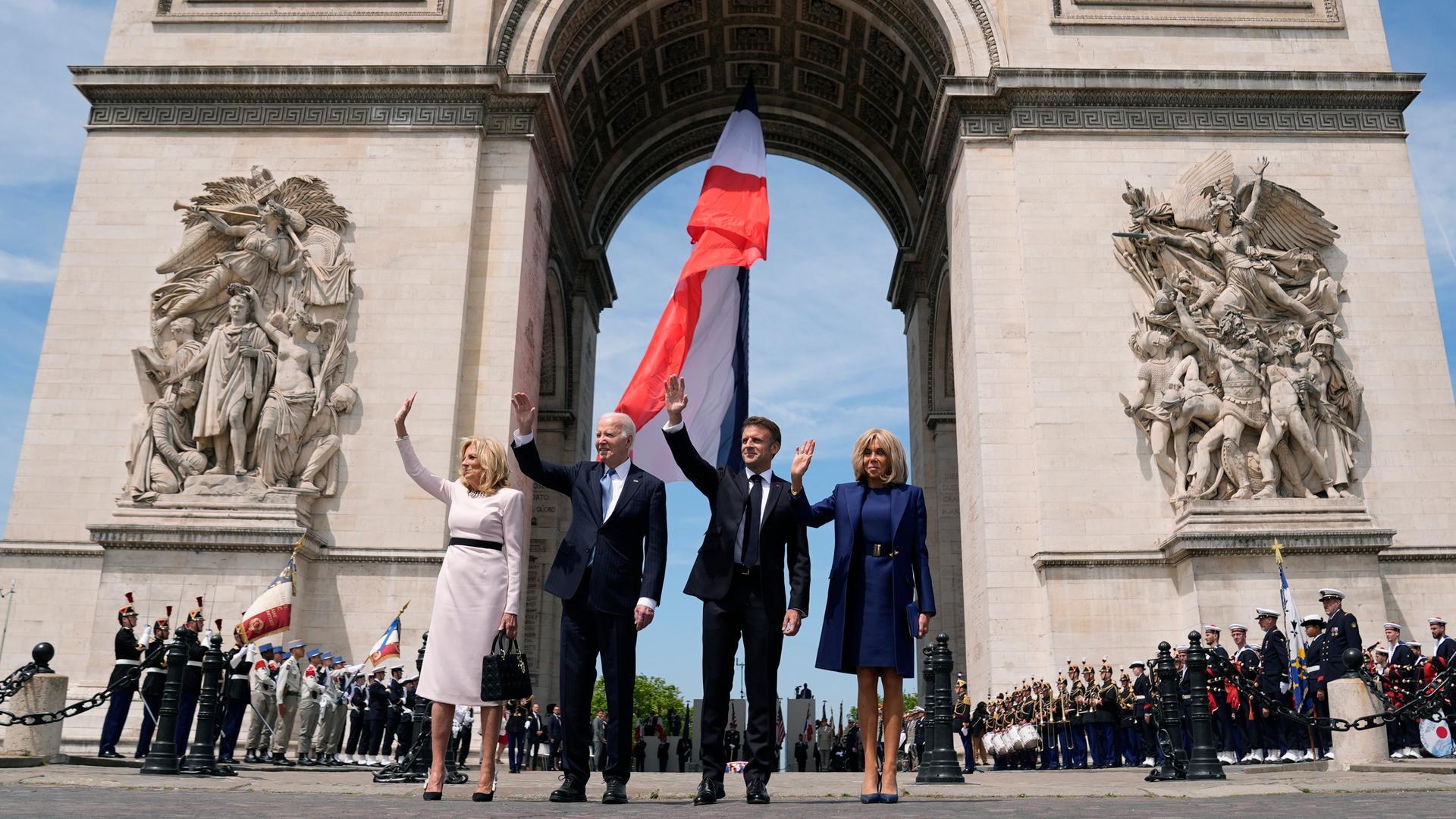 Vier Personen stehen winkend vor dem Arc de Triomphe in Paris: Jill Biden, First Lady der USA; Joe Biden, Präsident der USA; Emmanuel Macron, Präsident von Frankreich; und Brigitte Macron, First Lady von Frankreich - im Hintergrund ist die französische Flagge zu sehen