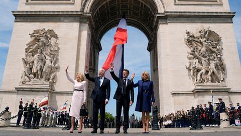 Vier Personen stehen winkend vor dem Arc de Triomphe in Paris: Jill Biden, First Lady der USA; Joe Biden, Präsident der USA; Emmanuel Macron, Präsident von Frankreich; und Brigitte Macron, First Lady von Frankreich - im Hintergrund ist die französische Flagge zu sehen