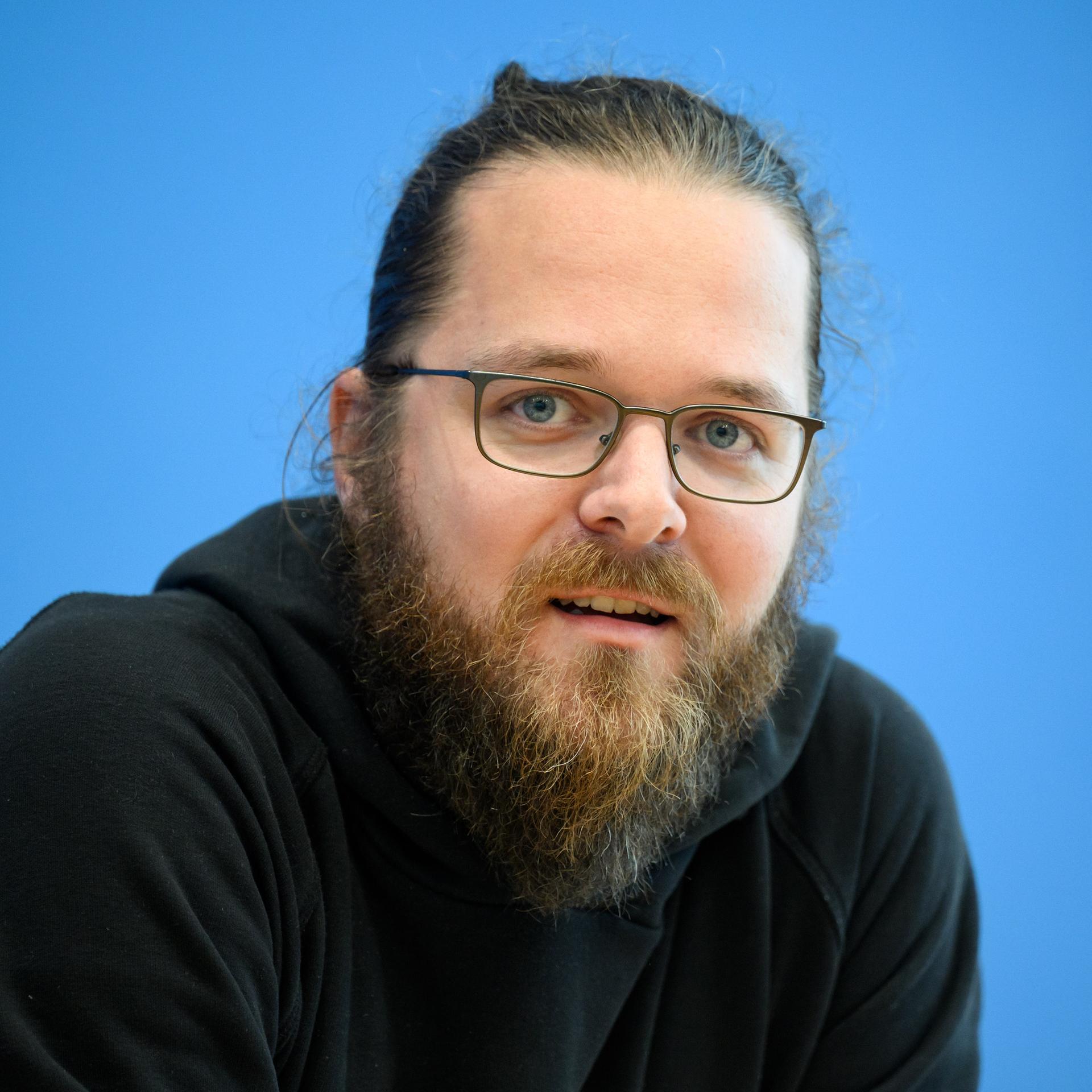 Arne Semsrott, Projektleiter von FragDenStaat, mit Bart, Dutt und Brille vor blauen Hintergrund.