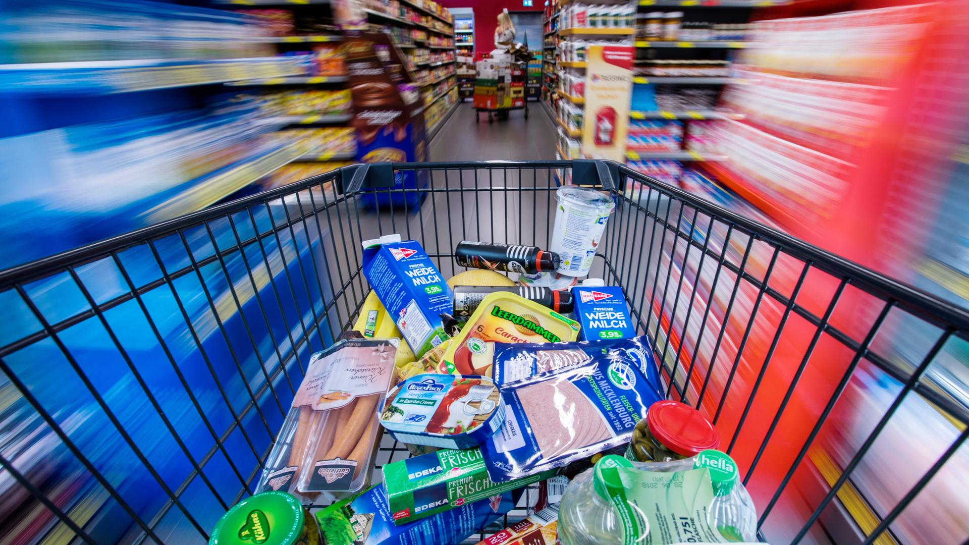 Ein Einkaufswagen mit Lebensmittel wird durch den Gang eines Supermarktes geschoben.