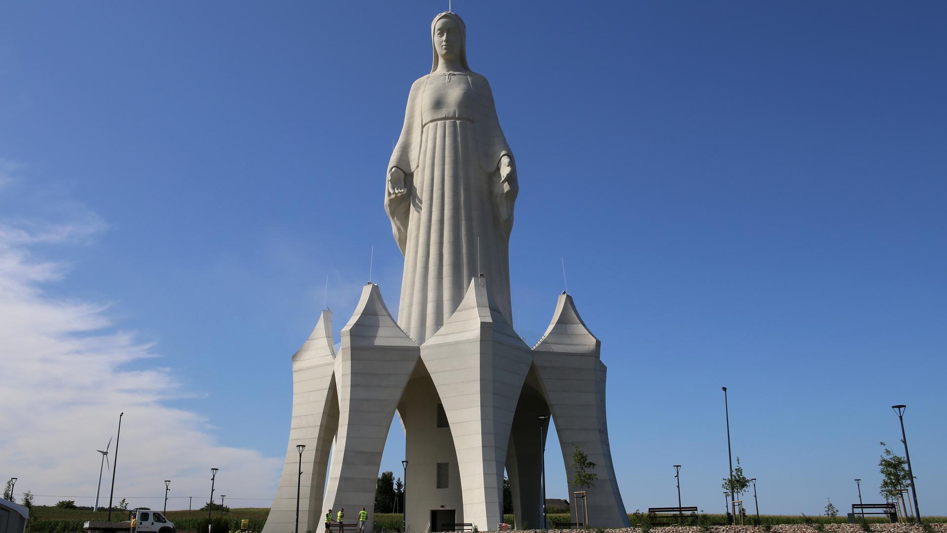 Eine riesige Marienstatue auf einem Feld. Sie steht auf einem hohen Sockel, in dem unten eine Tür ist. Die Menschen neben der Statue wirken winzig. Eine riesige Marienstatue auf einem Feld. Sie steht auf einem hohen Sockel, in dem unten eine Tür ist. Die Menschen neben der Statue wirken winzig.