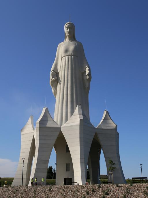 Eine riesige Marienstatue auf einem Feld. Sie steht auf einem hohen Sockel, in dem unten eine Tür ist. Die Menschen neben der Statue wirken winzig. 