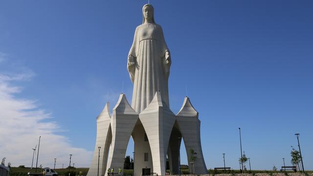 Eine riesige Marienstatue auf einem Feld. Sie steht auf einem hohen Sockel, in dem unten eine Tür ist. Die Menschen neben der Statue wirken winzig. 