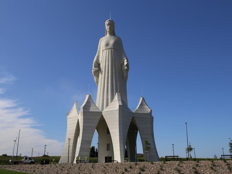 Eine riesige Marienstatue auf einem Feld. Sie steht auf einem hohen Sockel, in dem unten eine Tür ist. Die Menschen neben der Statue wirken winzig. 