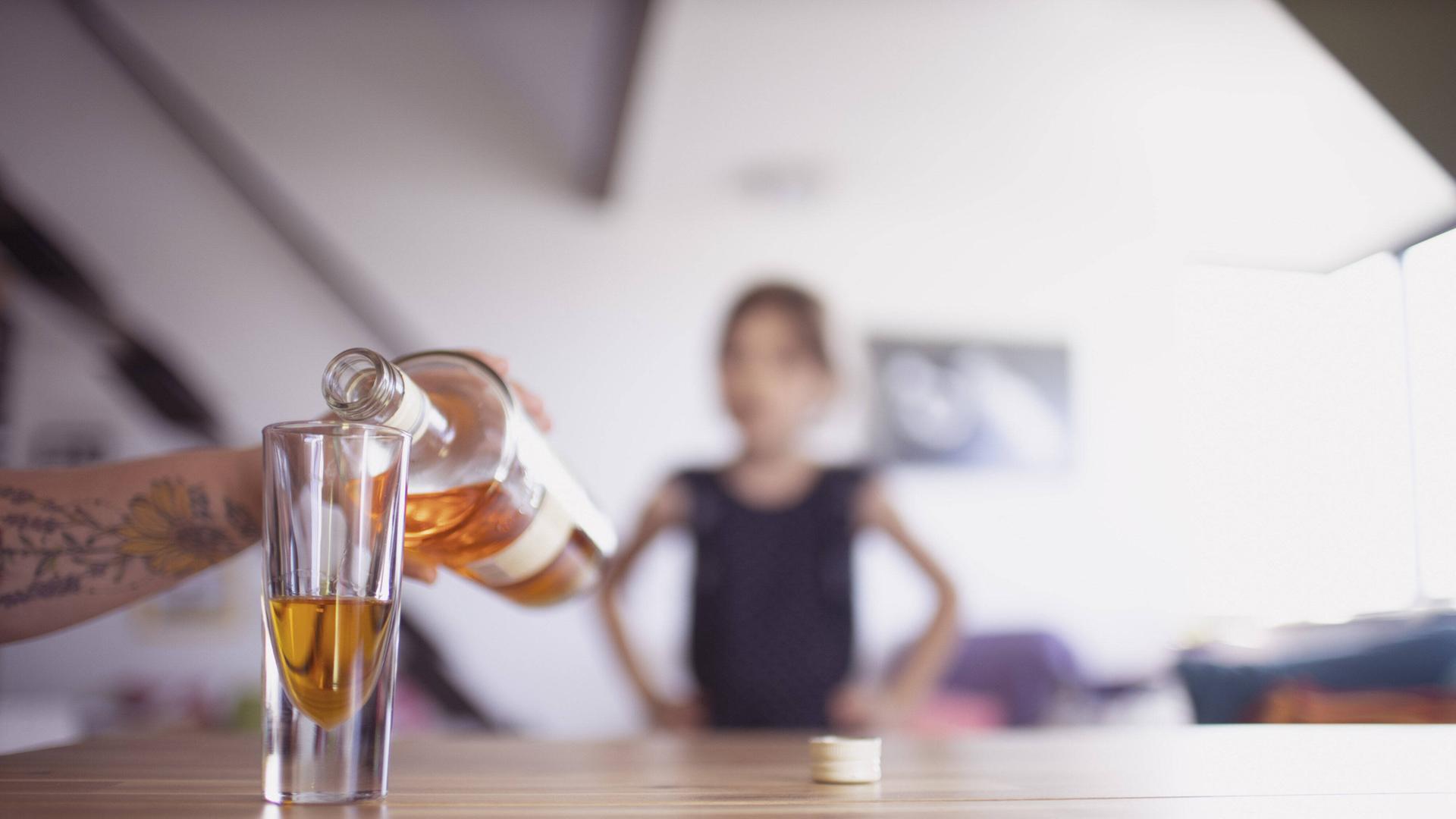 Hand gießt Whisky ins Glas, im Hintergrund ist unscharf ein Kind zu erkennen