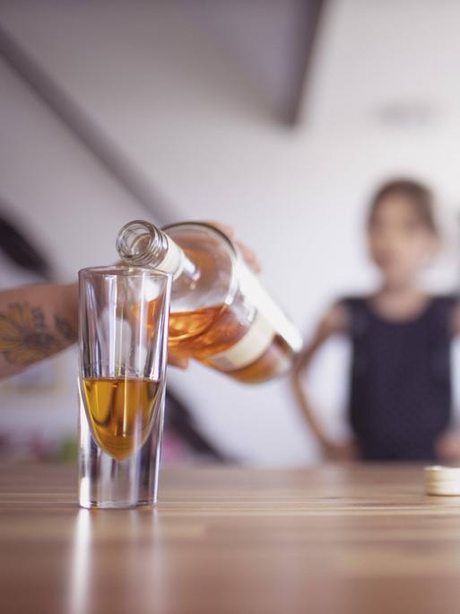 Hand gießt Whisky ins Glas, im Hintergrund ist unscharf ein Kind zu erkennen