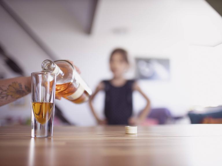 Hand gießt Whisky ins Glas, im Hintergrund ist unscharf ein Kind zu erkennen