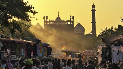 Menschenmenge auf einem belebten Markt bei Sonnenuntergang, im Hintergrund sind historische Gebäude sichtbar.