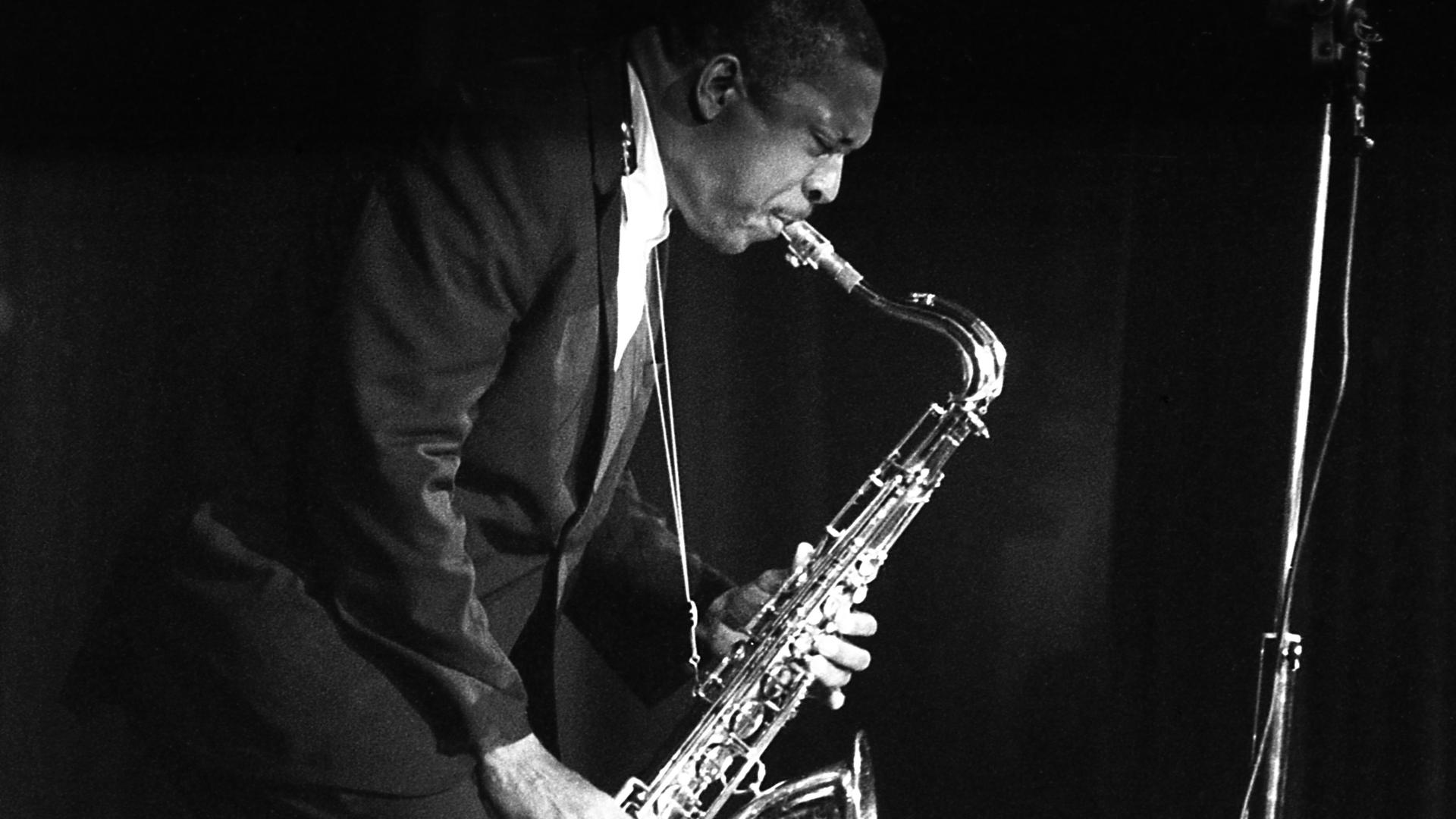 Historisches Schwarzweißfoto von John Coltrane, der auf einer Bühne steht und Saxophon spielt.